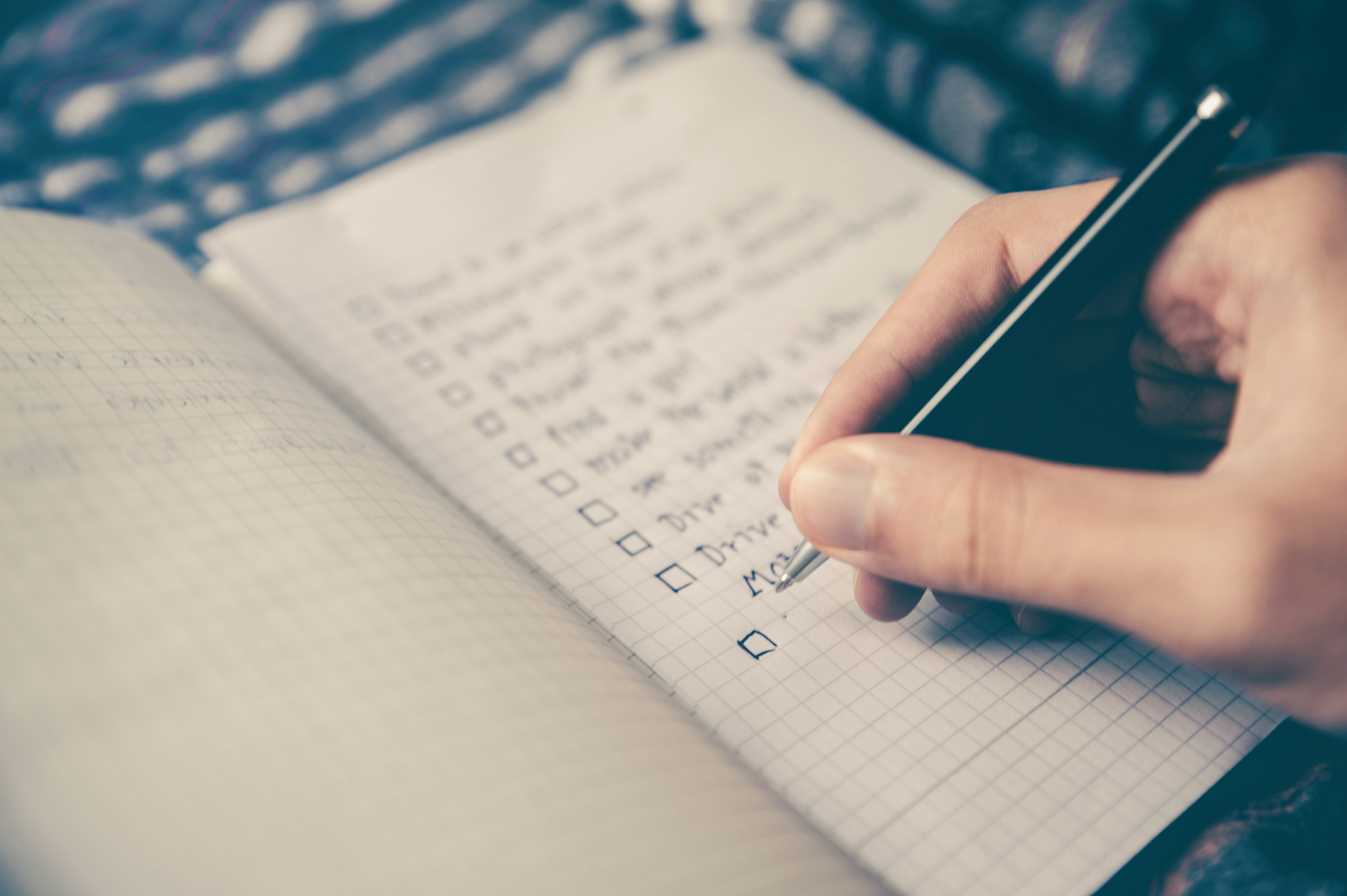 Een hand schrijft in een schriftje met ruitjes waarop reeds een lijst werd aangebracht die nu wordt afgevinkt. Achter de afbeelding zit een link naar een webpagina met meer informatie over de verschillende kosten die bij het huren van een woning komen kijken.