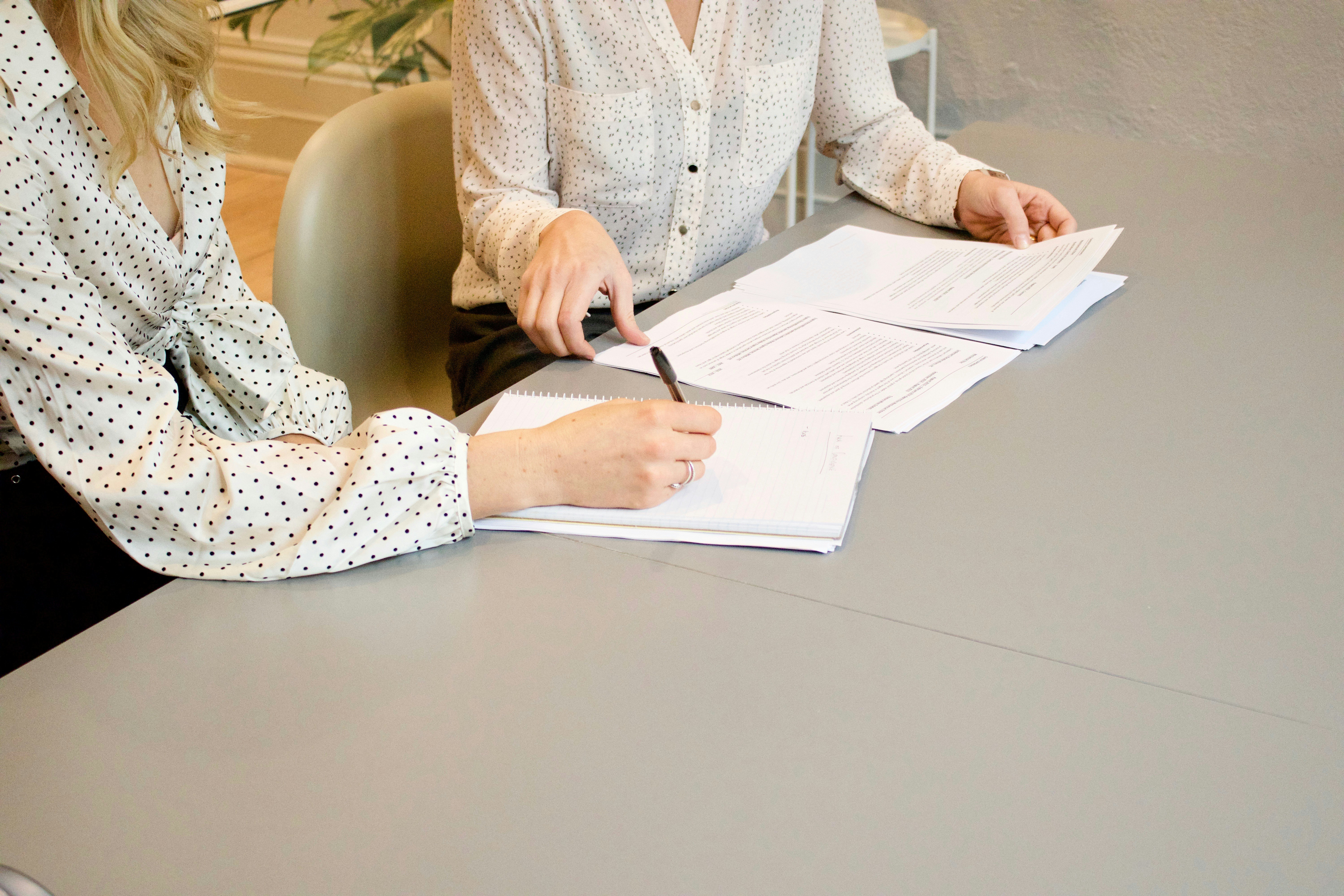 Twee vrouwen aan een vergadertafel bekijken documenten waarbij een van de twee de plekken aanduid waar de andere moet handtekenen.Achter de afbeelding zit een link naar een webpagina met meer informatie over het kiezen van een notariskantoor.